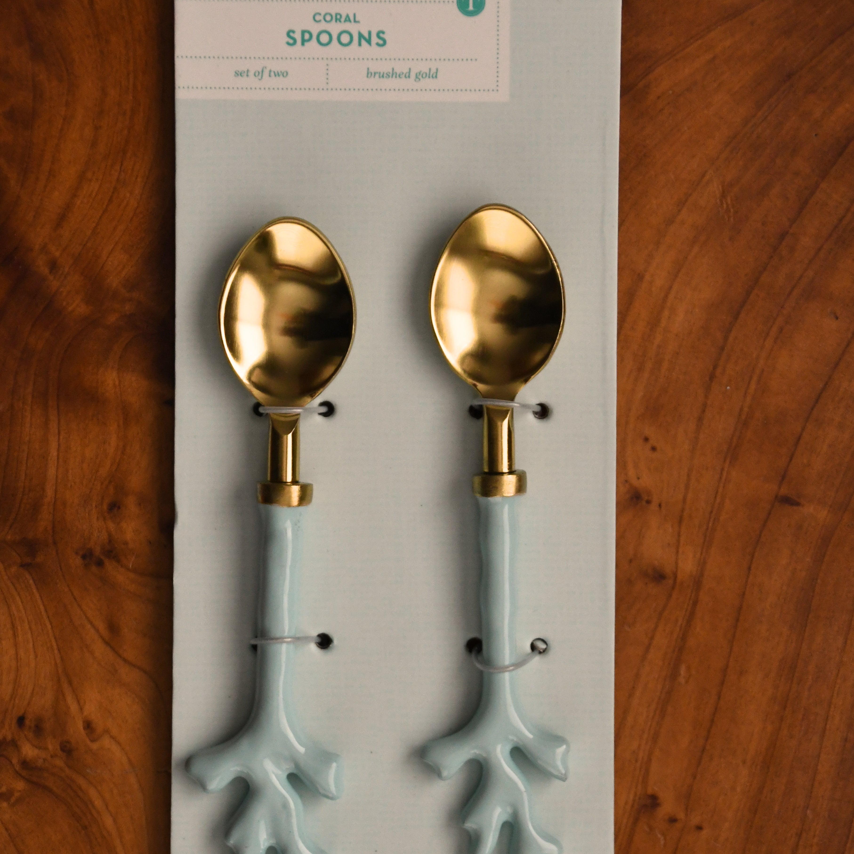 Two gold and light blue spoons on a Two's Company branded card against a wooden background