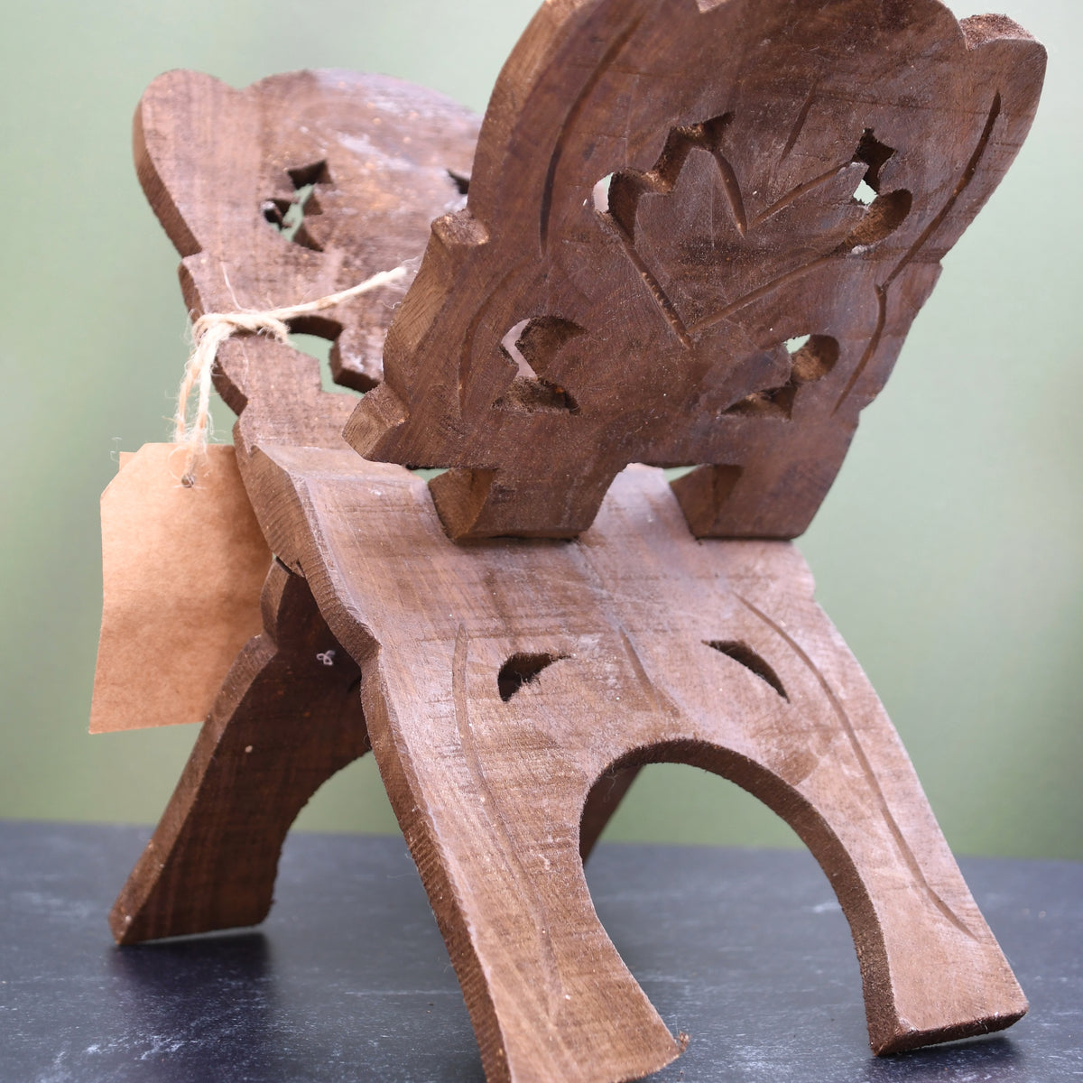 A small carved wooden chair or stool with a decorative backrest, photographed on a dark tabletop against a soft light green background. A small kraft paper tag is tied to the side.