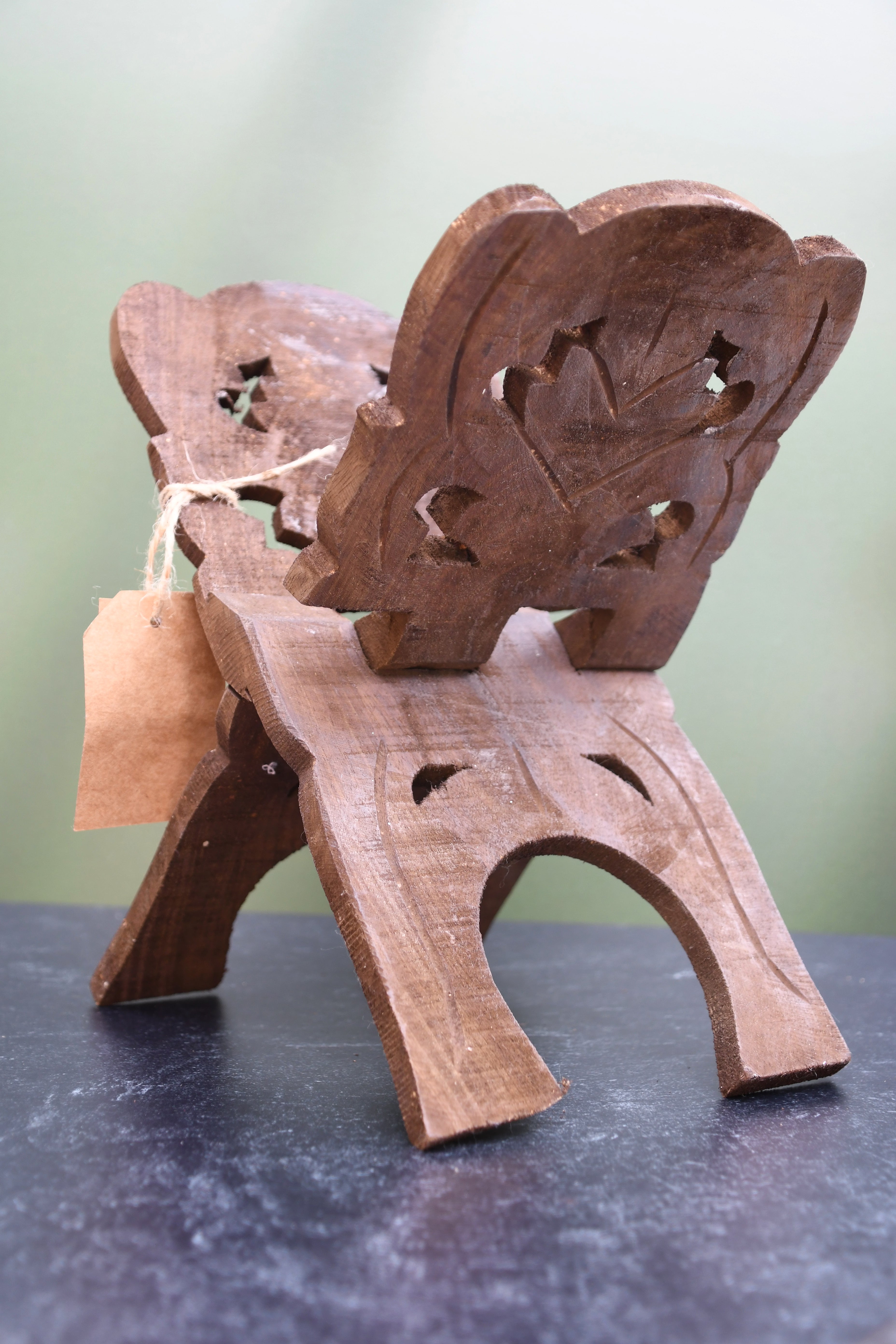 A small carved wooden chair or stool with a decorative backrest, photographed on a dark tabletop against a soft light green background. A small kraft paper tag is tied to the side.