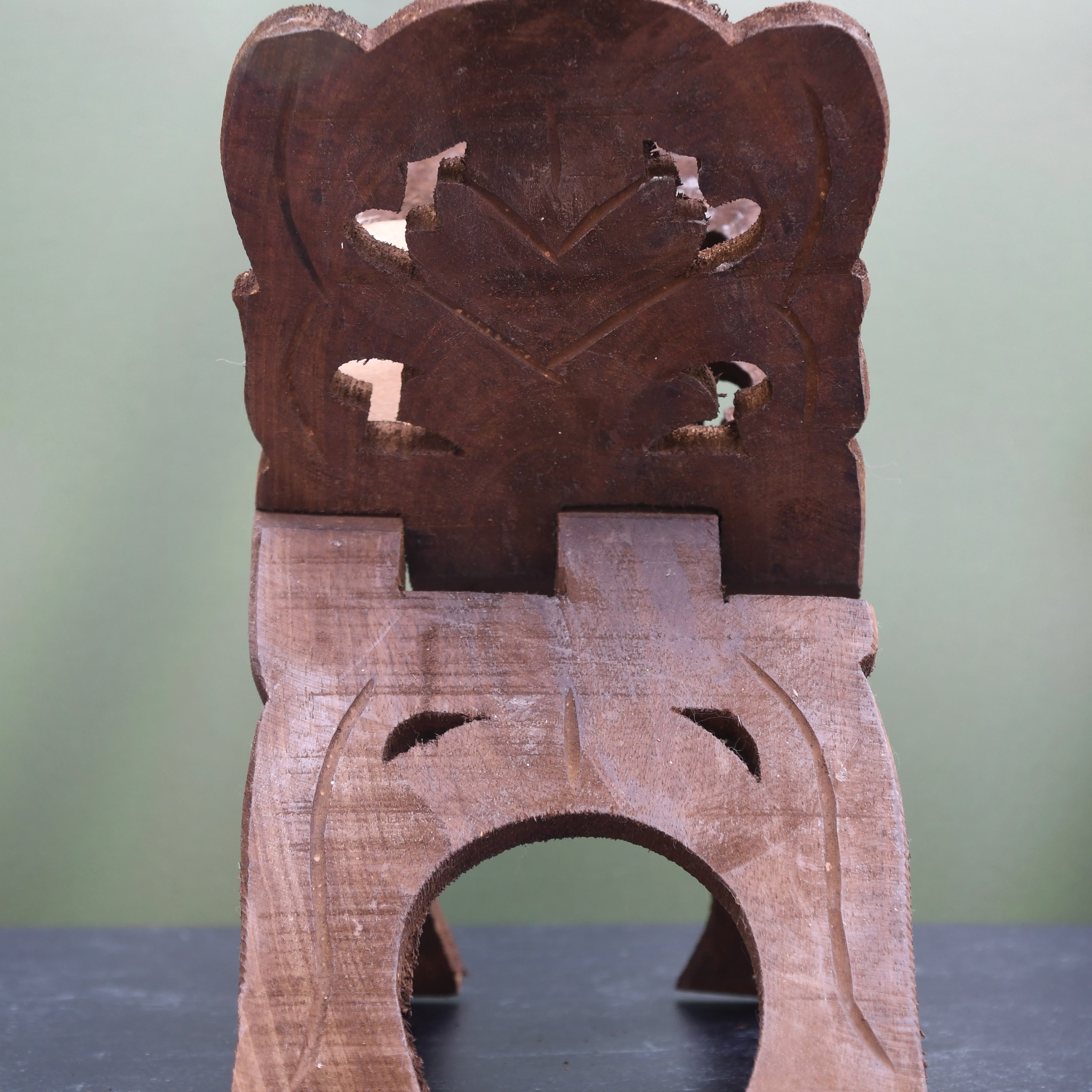 A small carved wooden stool or seat with a decorative cutout backrest and three legs, photographed on a dark tabletop against a soft light green background.