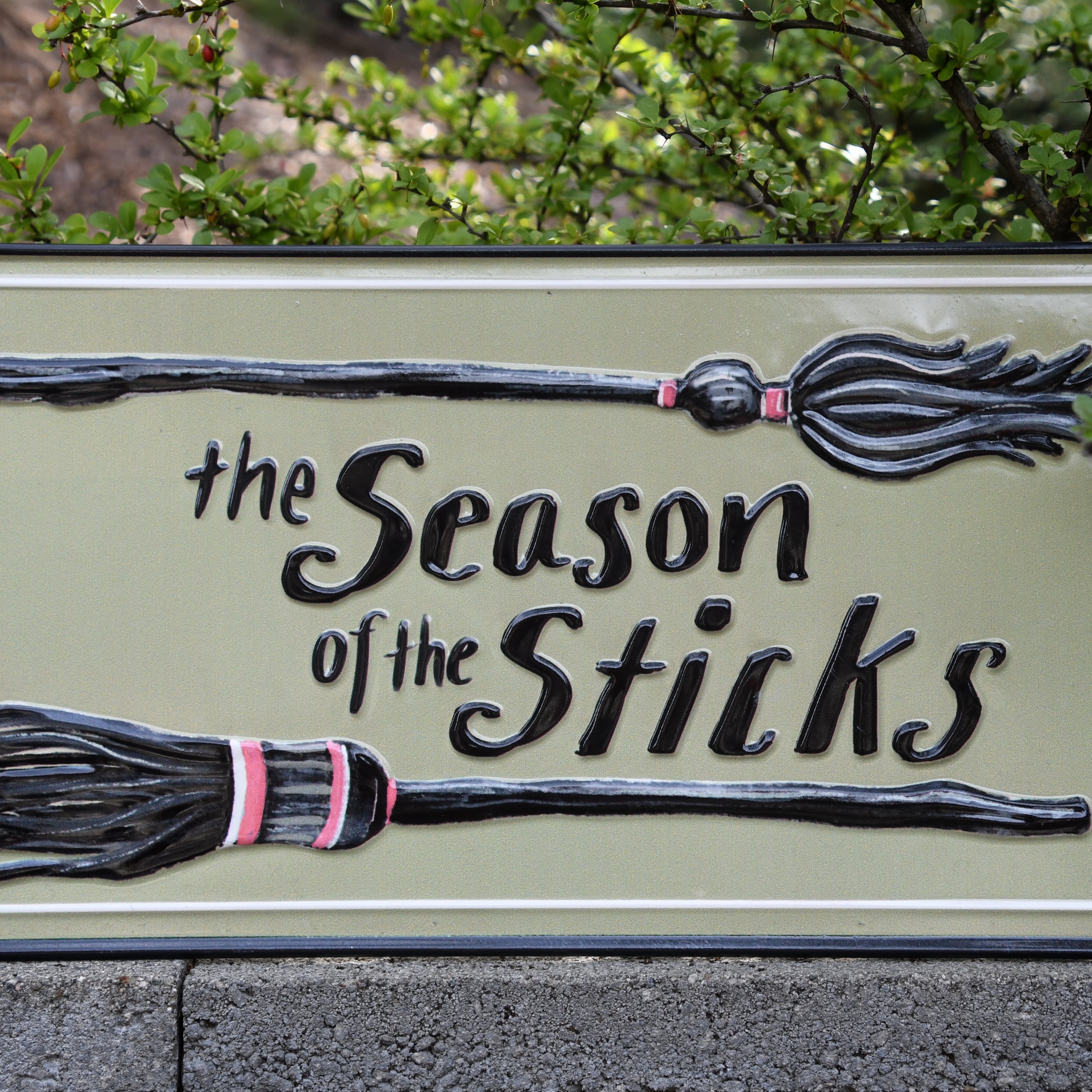 A decorative rectangular sign reading 'the Season of the Sticks' with two raised broomstick illustrations, photographed outdoors against green foliage and a stone wall.
