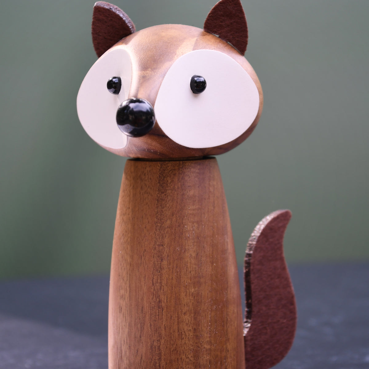 A wooden fox figurine with a smooth brown body, large white oval faces, black bead eyes and nose, and pointed ears, standing on a dark surface against a muted green background.