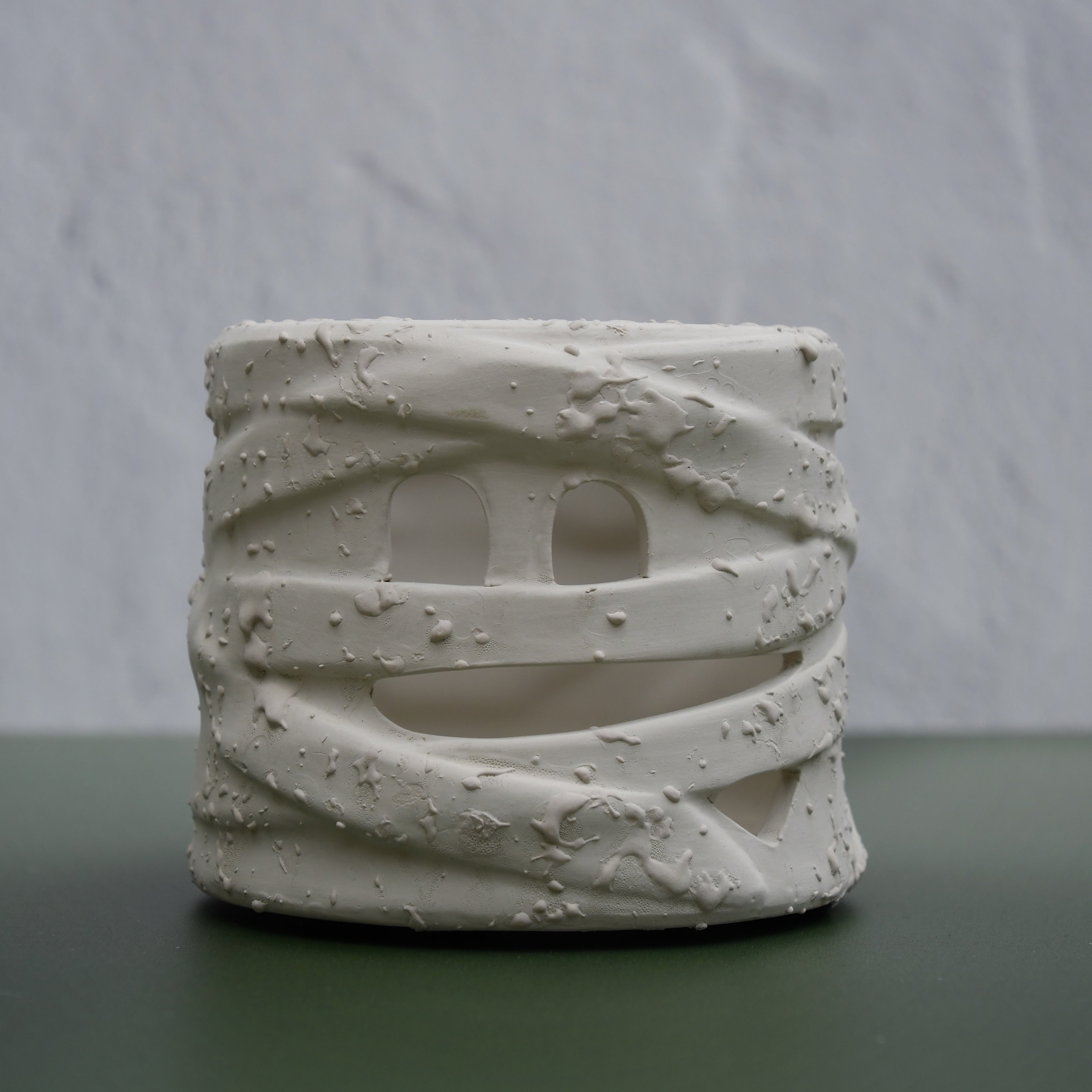 A small cylindrical white ceramic planter with a textured, wrapped design and cutout details, displayed on a dark green surface against a light gray textured wall.