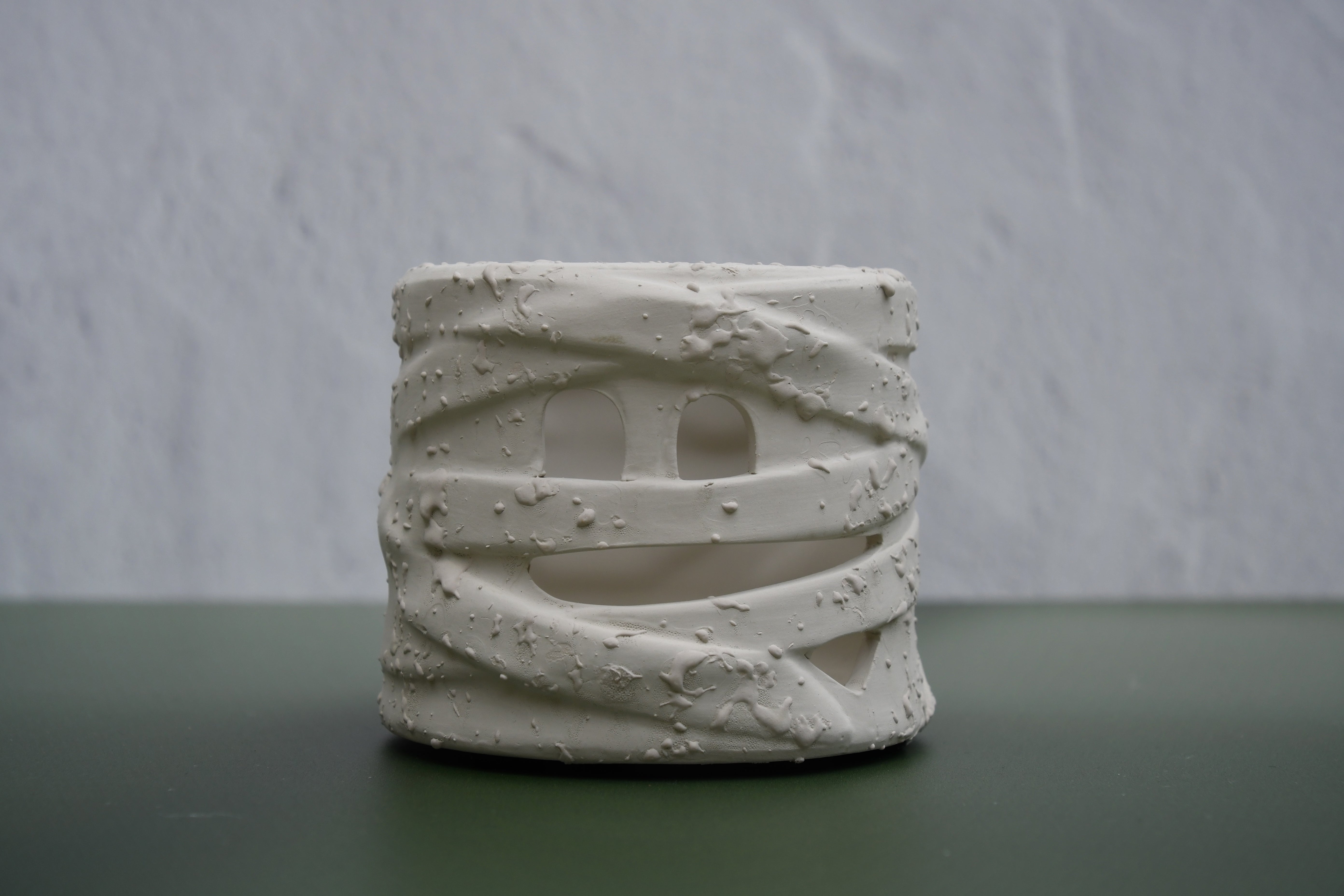 A small cylindrical white ceramic planter with a textured, wrapped design and cutout details, displayed on a dark green surface against a light gray textured wall.