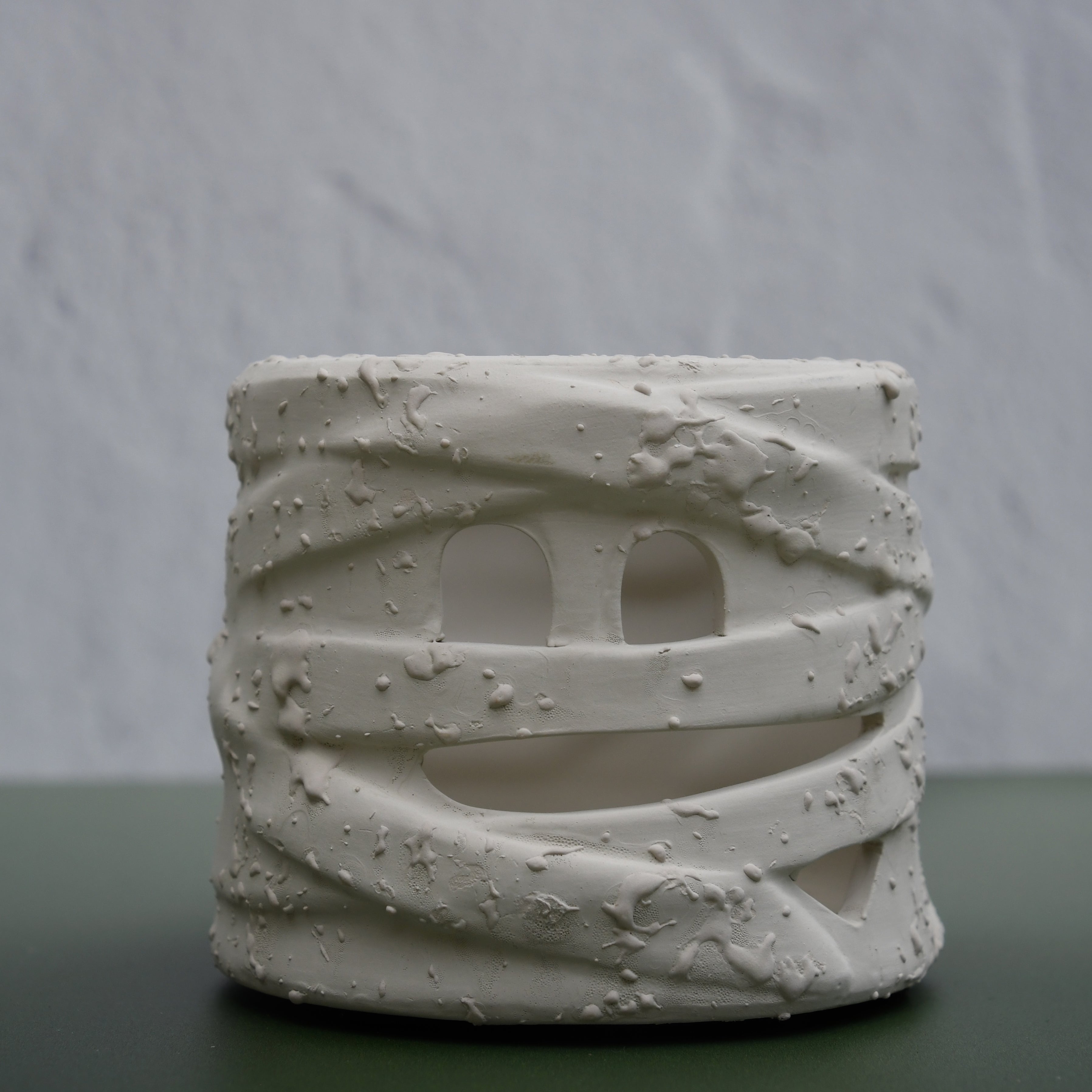 A small cylindrical ceramic candle holder with a textured, wrapped mummy-style design and cutout eyes, displayed on a dark green surface against a light gray textured background.