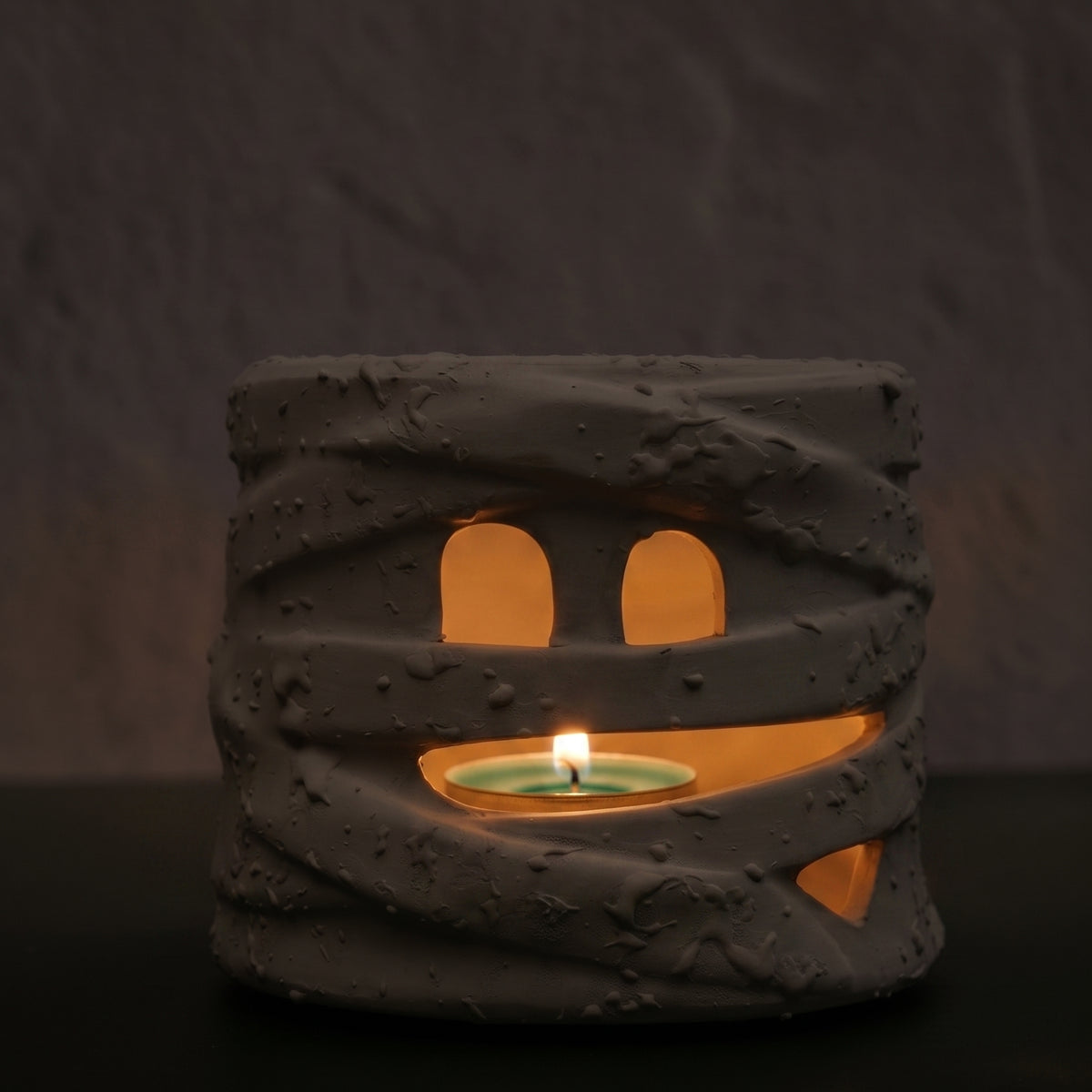 A mummy-themed candle holder and tea light holder made of textured gray resin, featuring cutout eyes and a mouth that reveal a glowing green tea light inside, set against a dark textured background.