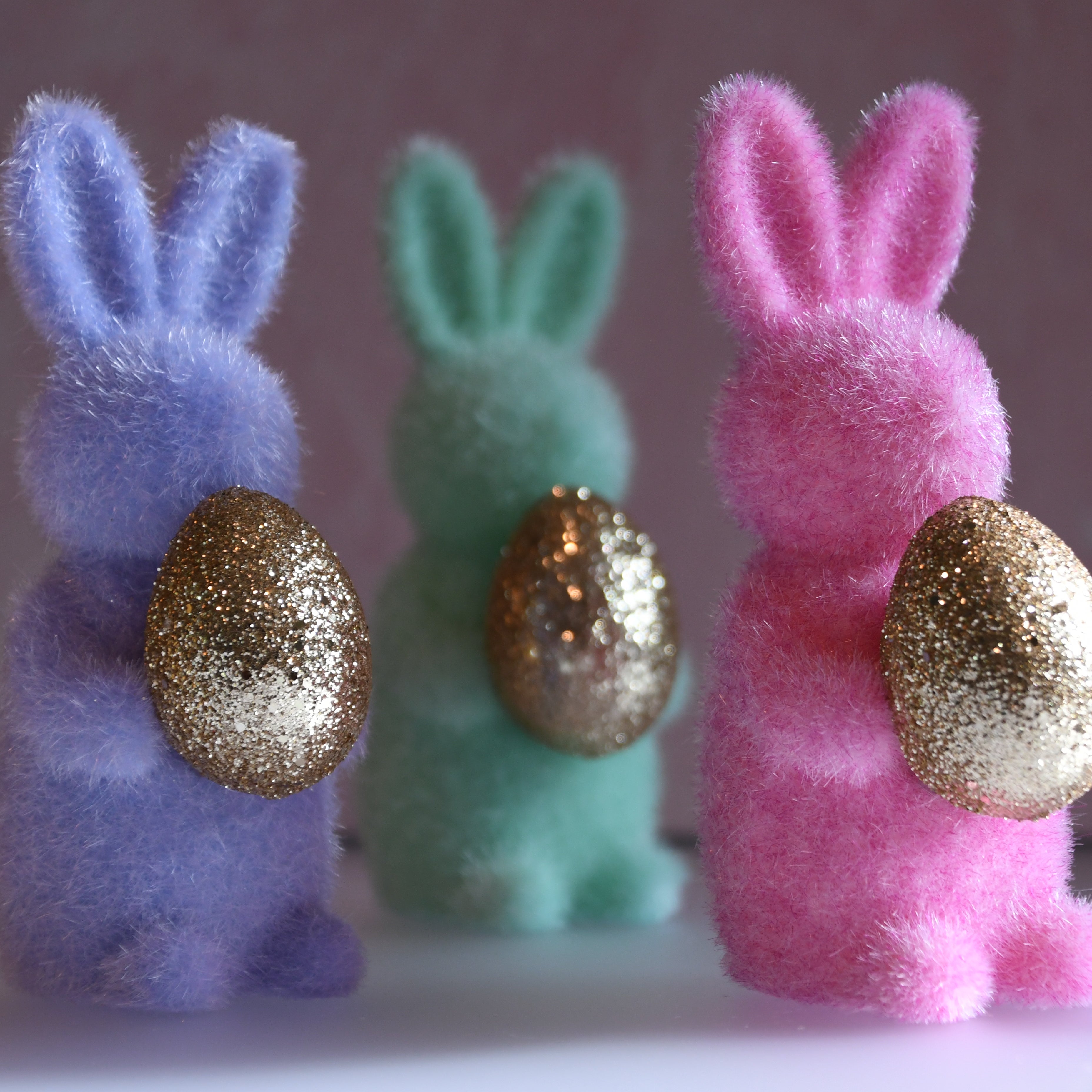 Three colorful bunny figurines with glittery eggs on a plain background