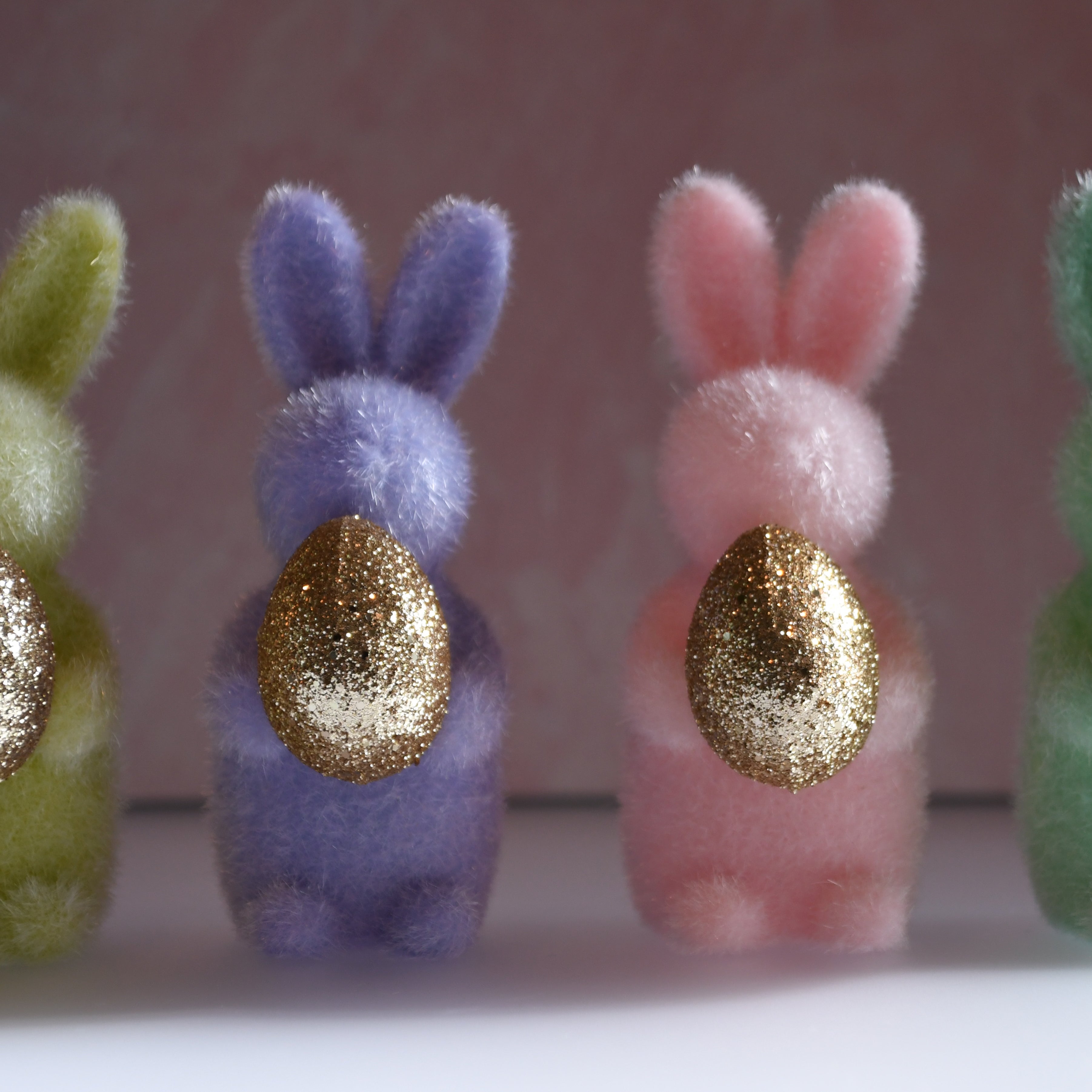 Four colorful felt bunnies with gold eggs on a white surface and light pink background