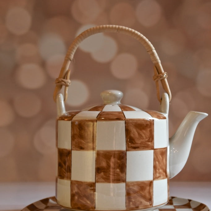 Checkered ceramic teapot with wicker handle on a matching tray against a blurred background