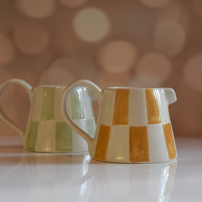 Two ceramic pitchers with checkered patterns on a reflective surface with a blurred bokeh background.
