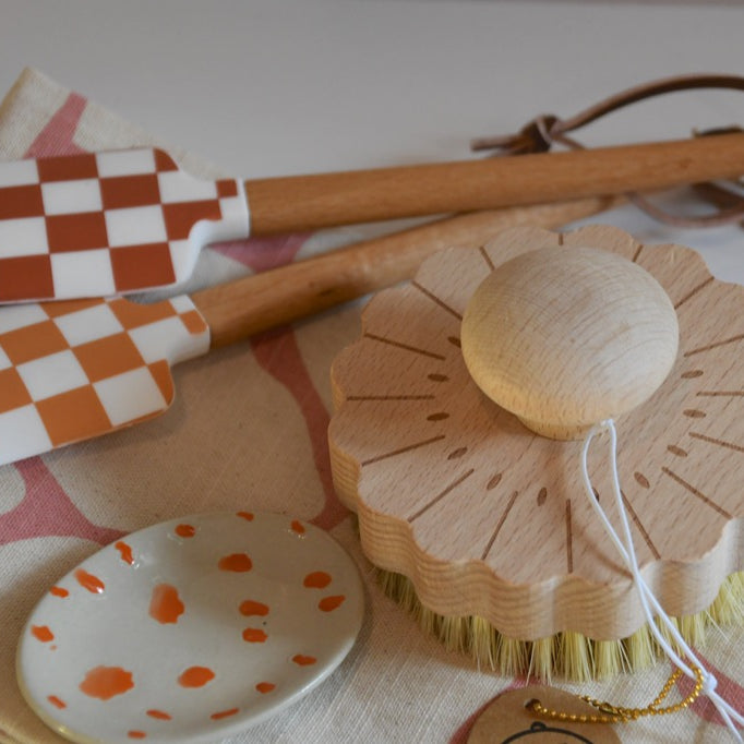 Set of kitchen utensils including checkered spatulas, a wooden brush, and a polka dot dish on a patterned cloth.