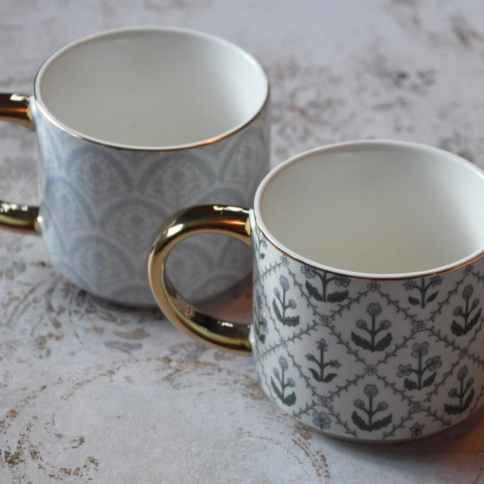 Two patterned mugs with gold handles on a textured surface