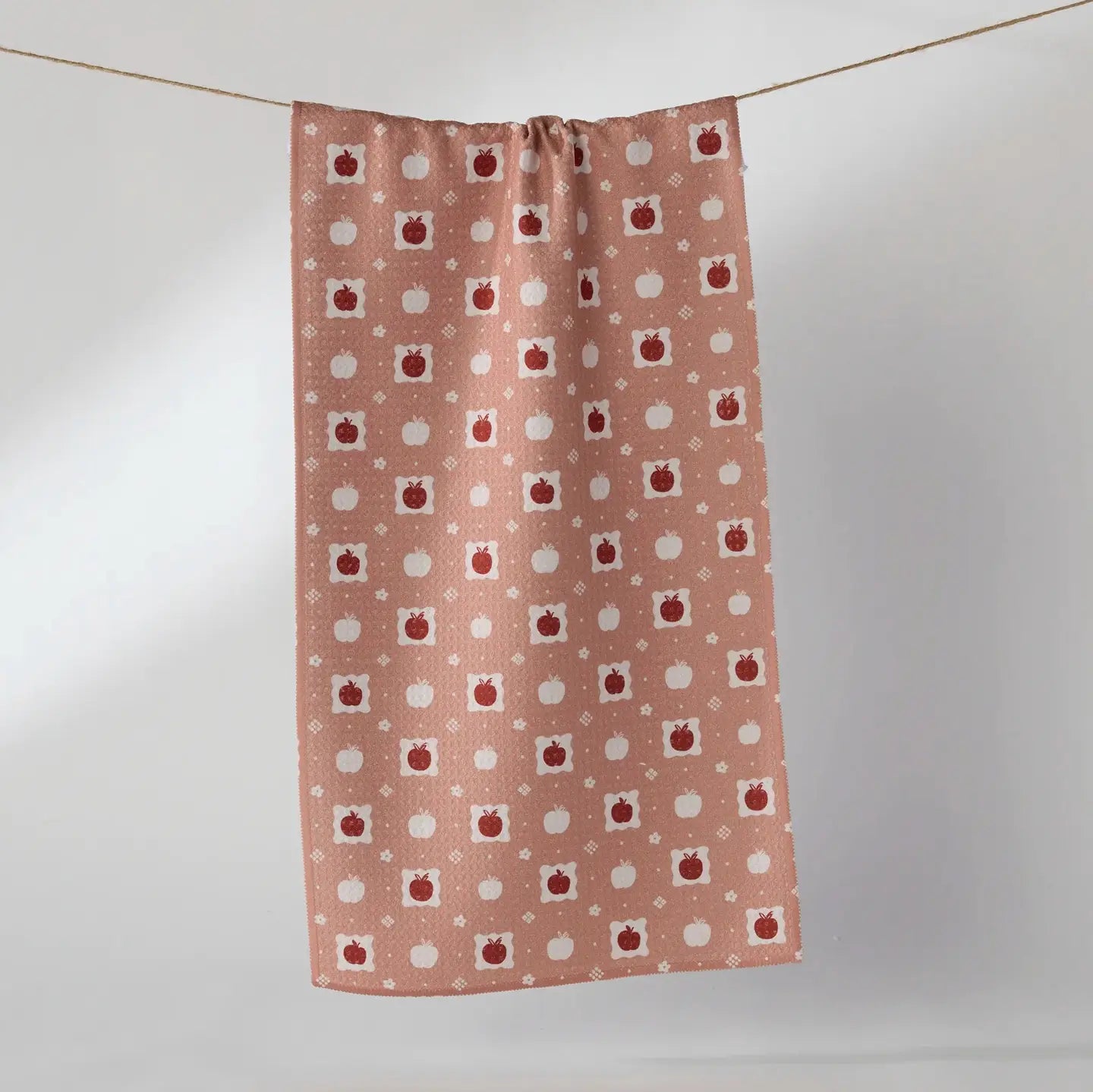 A rectangular dust cloth or curtain with a repeating pattern of red apples and white pumpkins on a dusty pink background, hanging from a string against a light gray wall.