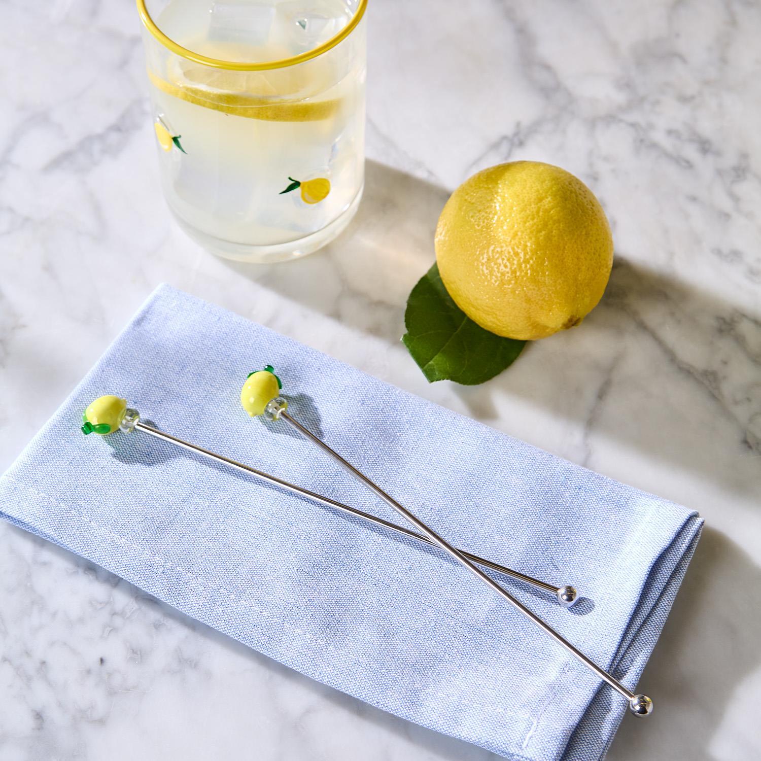 Two lemon-themed cocktail stirrers on a blue napkin with a glass of lemonade and a lemon in the background.