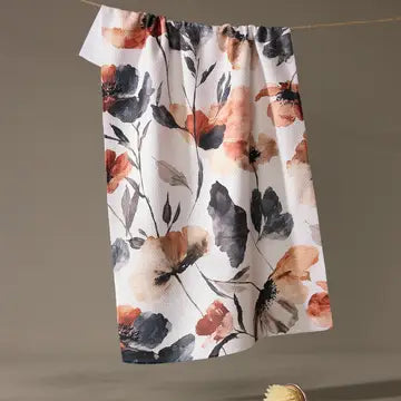 A white kitchen towel with a watercolor-style floral print in orange, black, and gray, hanging from a string against a neutral gray background.