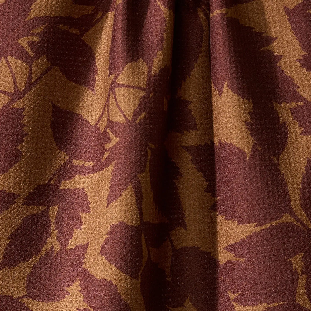 A close-up of draped fabric with a waffle-weave texture, featuring a dark burgundy leaf pattern over a warm tan background.