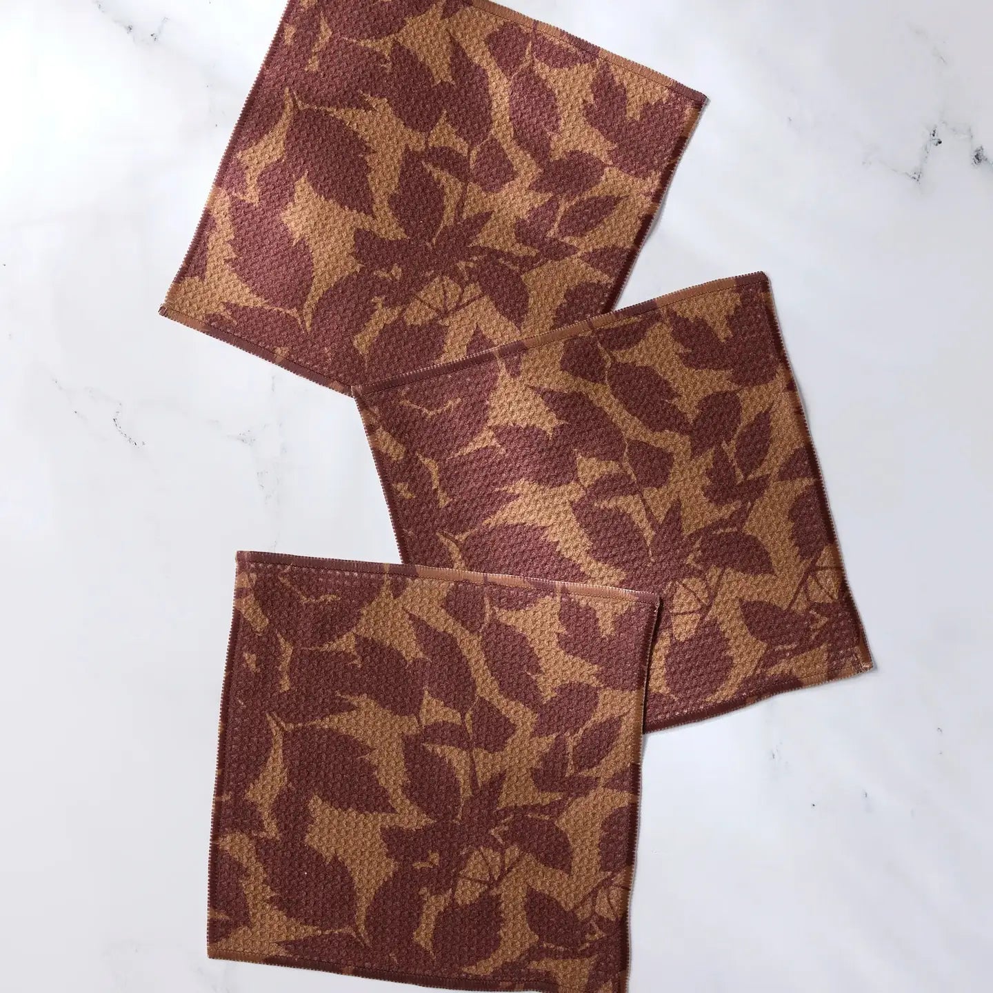 Three square woven coasters with a brown and tan leaf pattern, arranged diagonally on a white marble surface.
