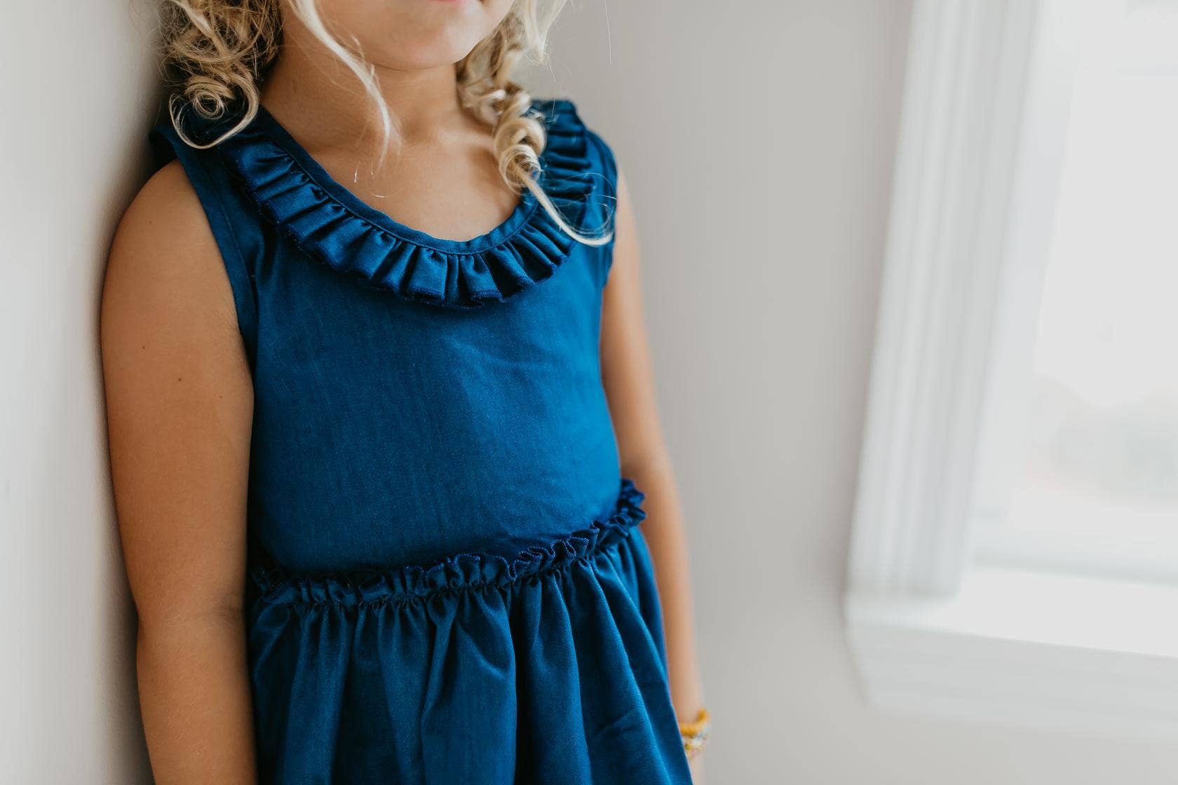 This sleeveless blue denim dress features a playful ruffle neckline and intricate lace hemline