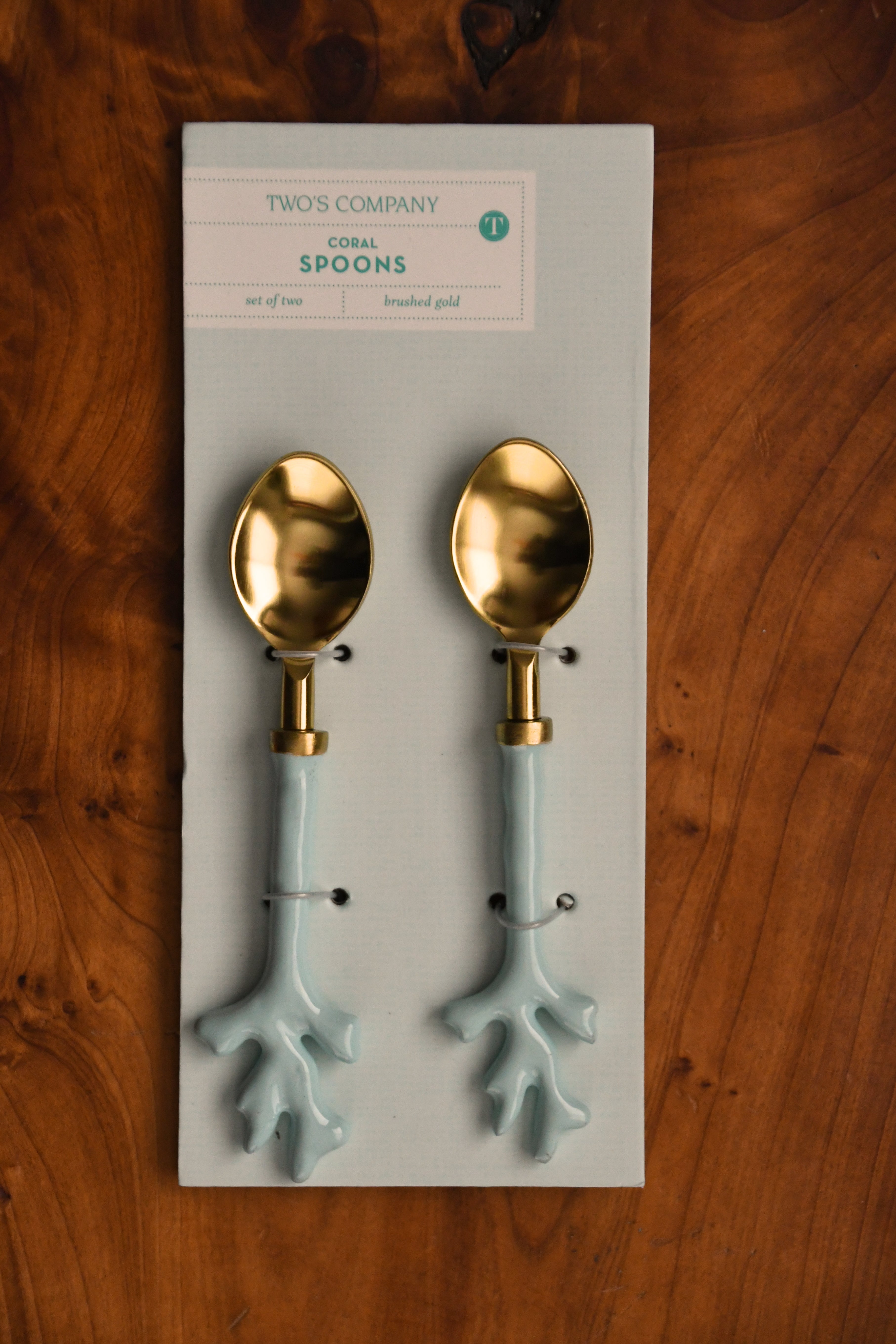 Two gold and light blue spoons on a Two's Company branded card against a wooden background