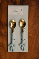 Two gold and light blue spoons on a Two's Company branded card against a wooden background