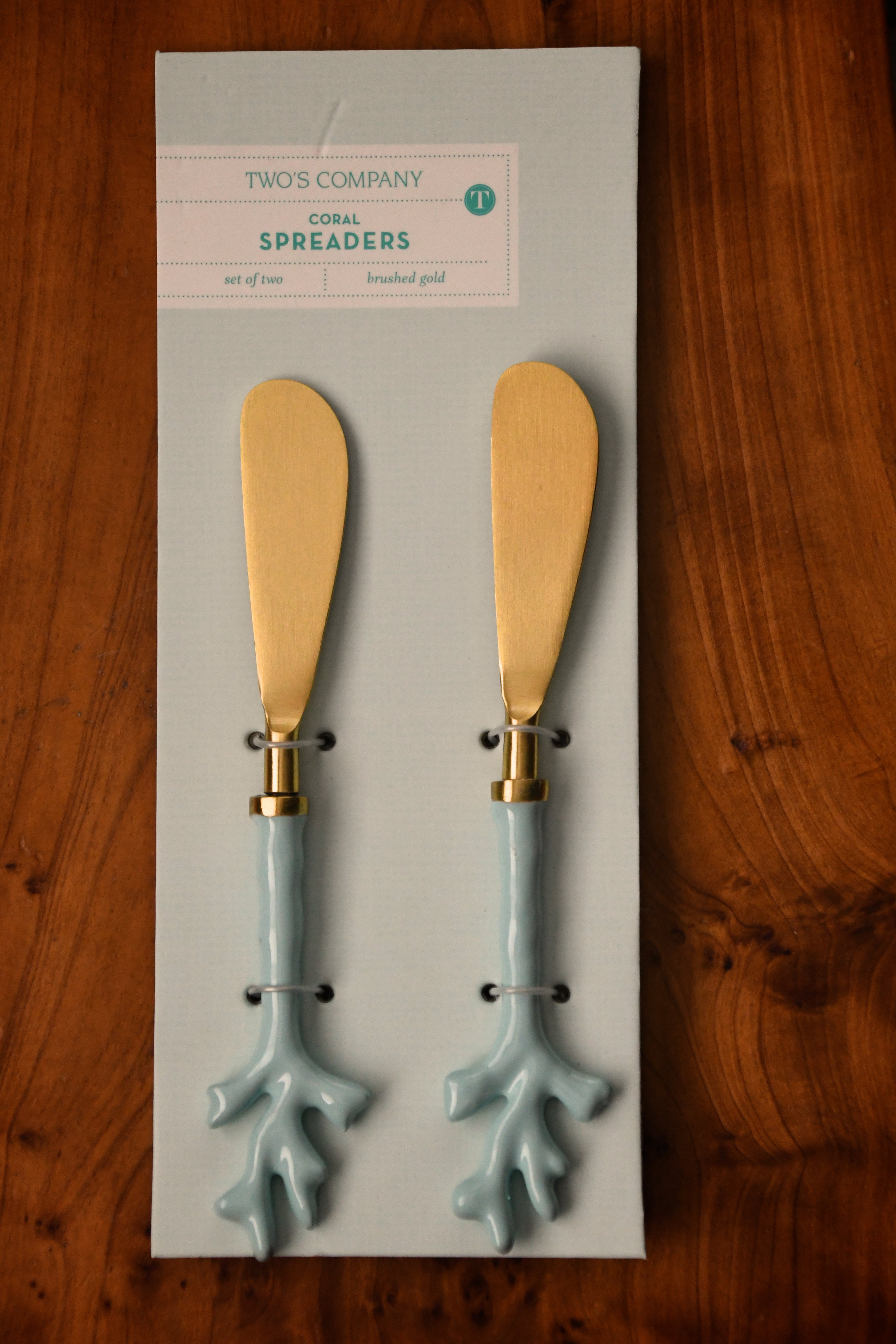 Two coral spreaders on a Two's Company branded card against a wooden background