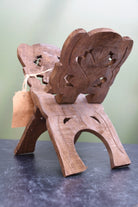 A small carved wooden chair or stool with a decorative backrest, photographed on a dark tabletop against a soft light green background. A small kraft paper tag is tied to the side.