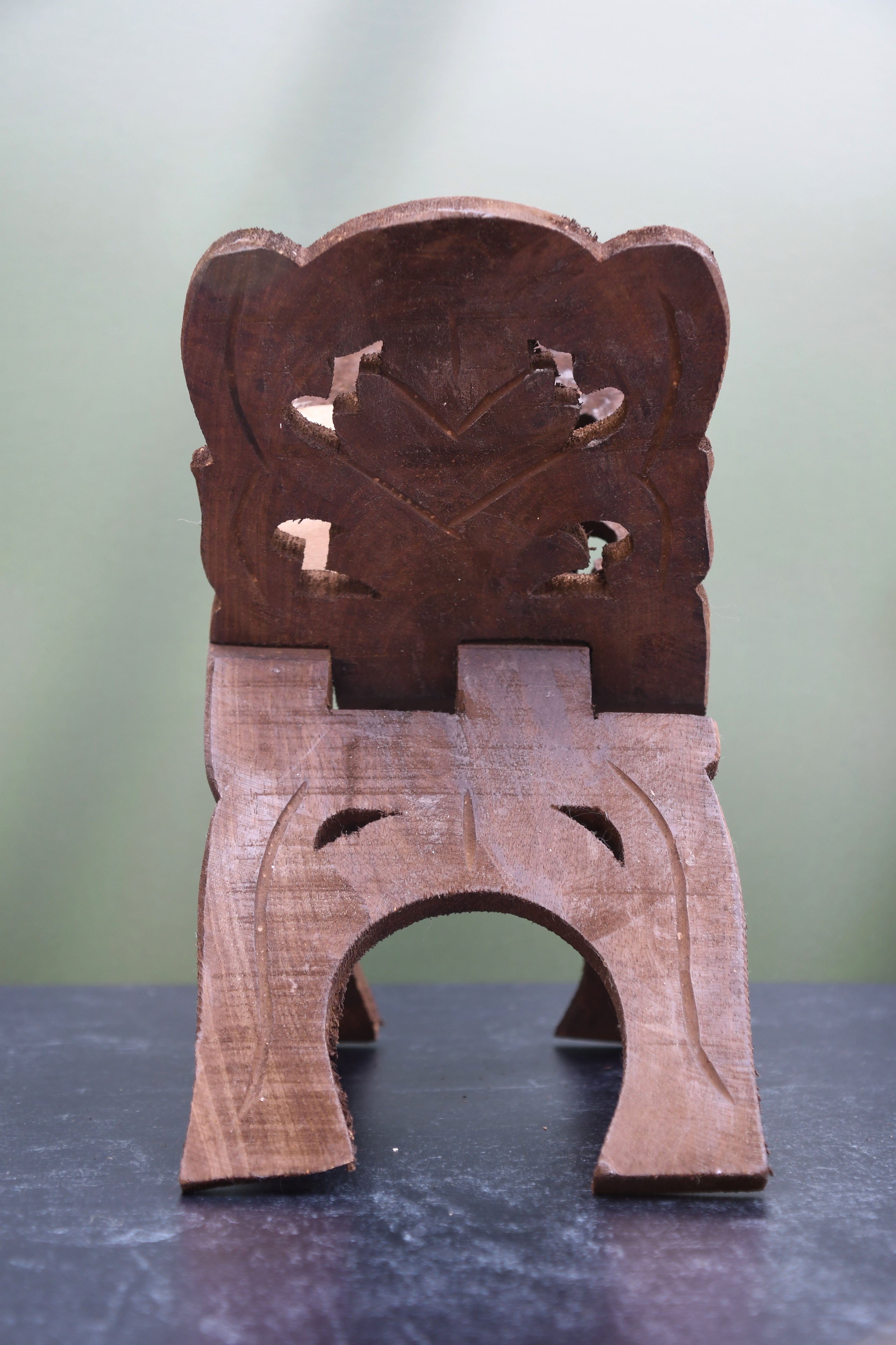 A small carved wooden stool or seat with a decorative cutout backrest and three legs, photographed on a dark tabletop against a soft light green background.