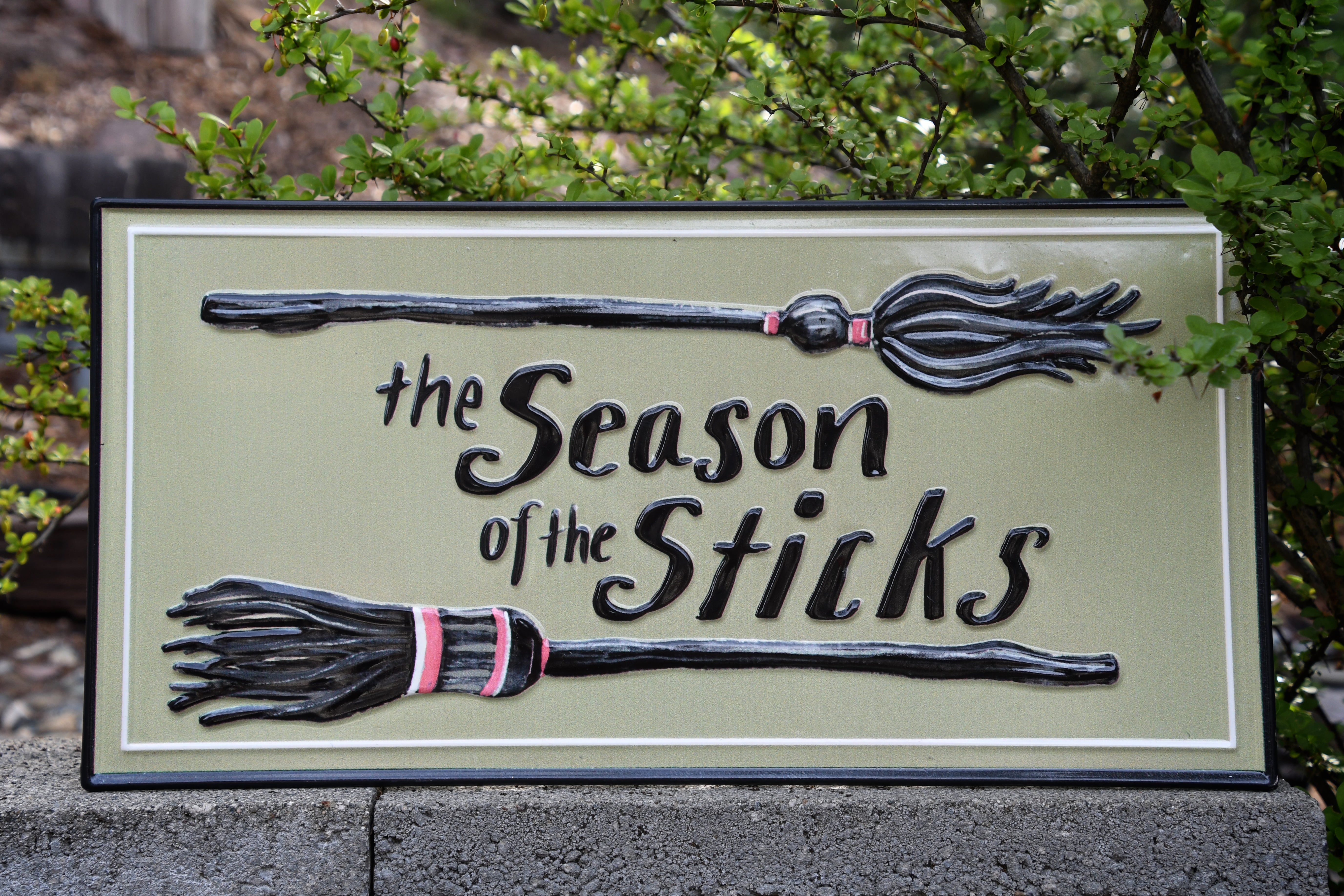 A decorative rectangular sign reading 'the Season of the Sticks' with two raised broomstick illustrations, photographed outdoors against green foliage and a stone wall.
