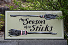 A decorative rectangular sign reading 'the Season of the Sticks' with two raised broomstick illustrations, photographed outdoors against green foliage and a stone wall.