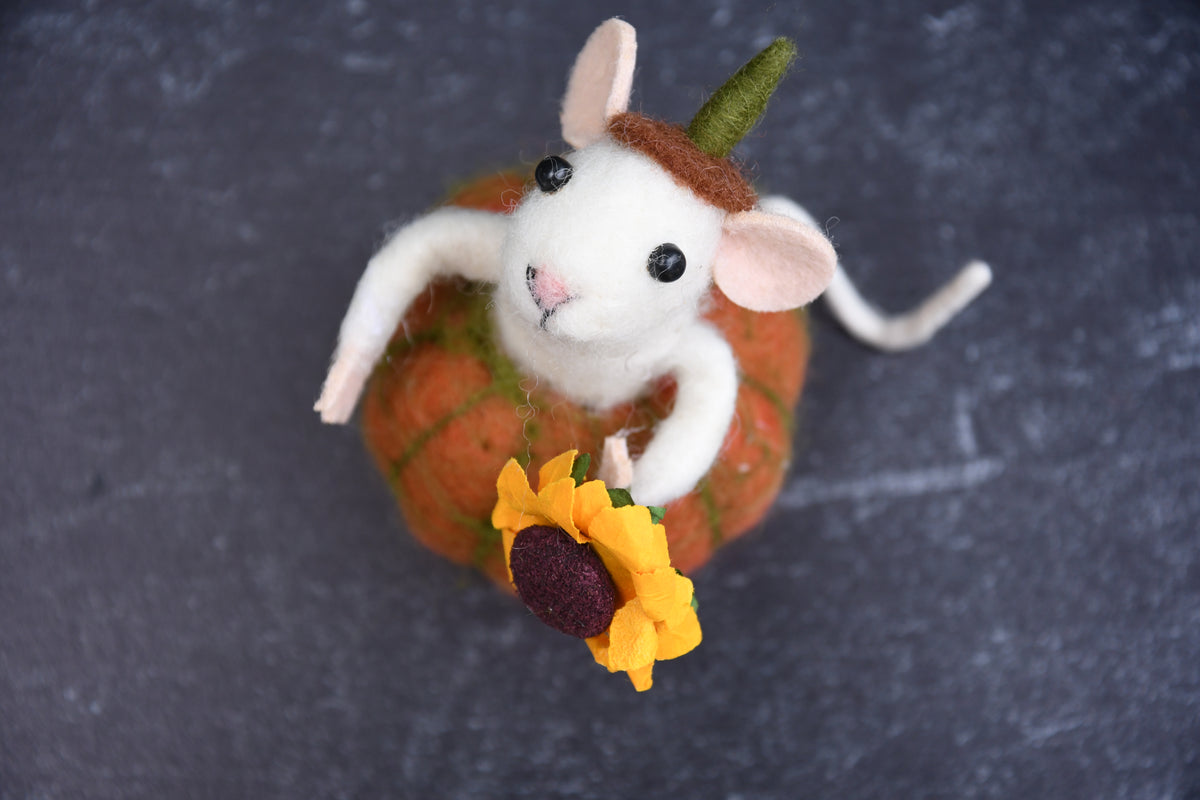 A handcrafted felted wool art piece of a white mouse with pink ears and a green hat, sitting on top of an orange pumpkin and holding a yellow sunflower, set against a dark slate background.