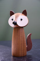 A wooden fox figurine with a smooth brown body, large white oval faces, black bead eyes and nose, and pointed ears, standing on a dark surface against a muted green background.