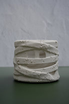 A small cylindrical ceramic candle holder with a textured, wrapped mummy-style design and cutout eyes, displayed on a dark green surface against a light gray textured background.
