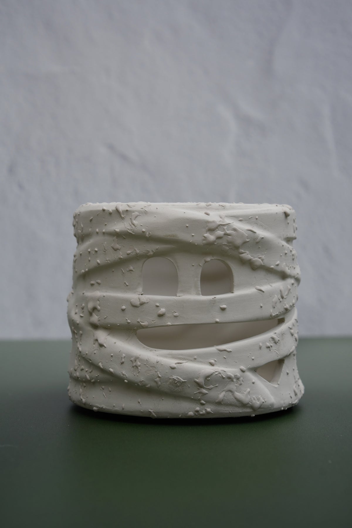 A small cylindrical ceramic candle holder with a textured, wrapped mummy-style design and cutout eyes, displayed on a dark green surface against a light gray textured background.