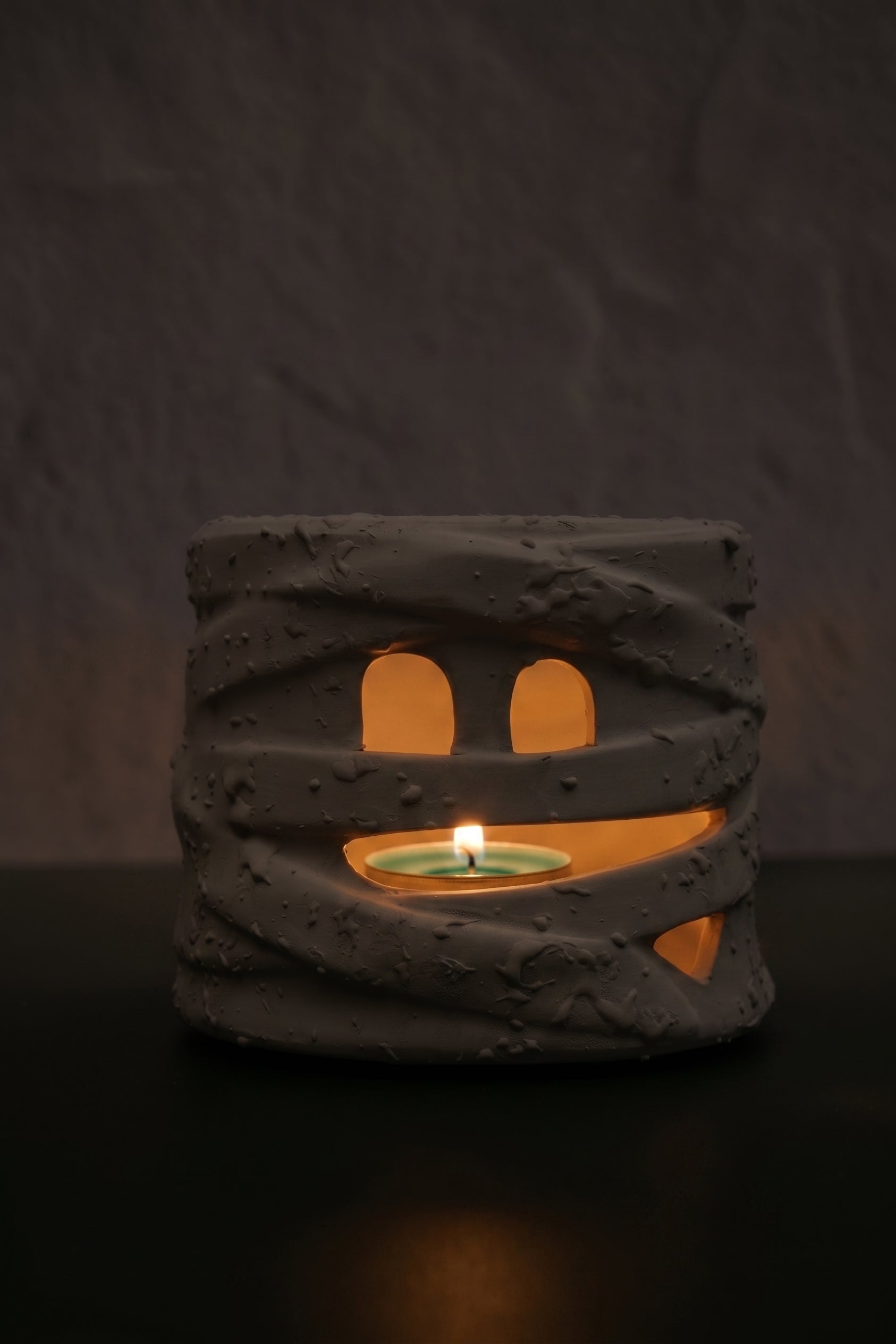 A mummy-themed candle holder and tea light holder made of textured gray resin, featuring cutout eyes and a mouth that reveal a glowing green tea light inside, set against a dark textured background.