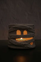 A mummy-themed candle holder and tea light holder made of textured gray resin, featuring cutout eyes and a mouth that reveal a glowing green tea light inside, set against a dark textured background.