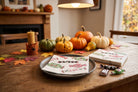 A styled autumn table scene with small pumpkins and fall leaves on a wooden table, featuring printed napkins and candy props in a warm indoor setting.