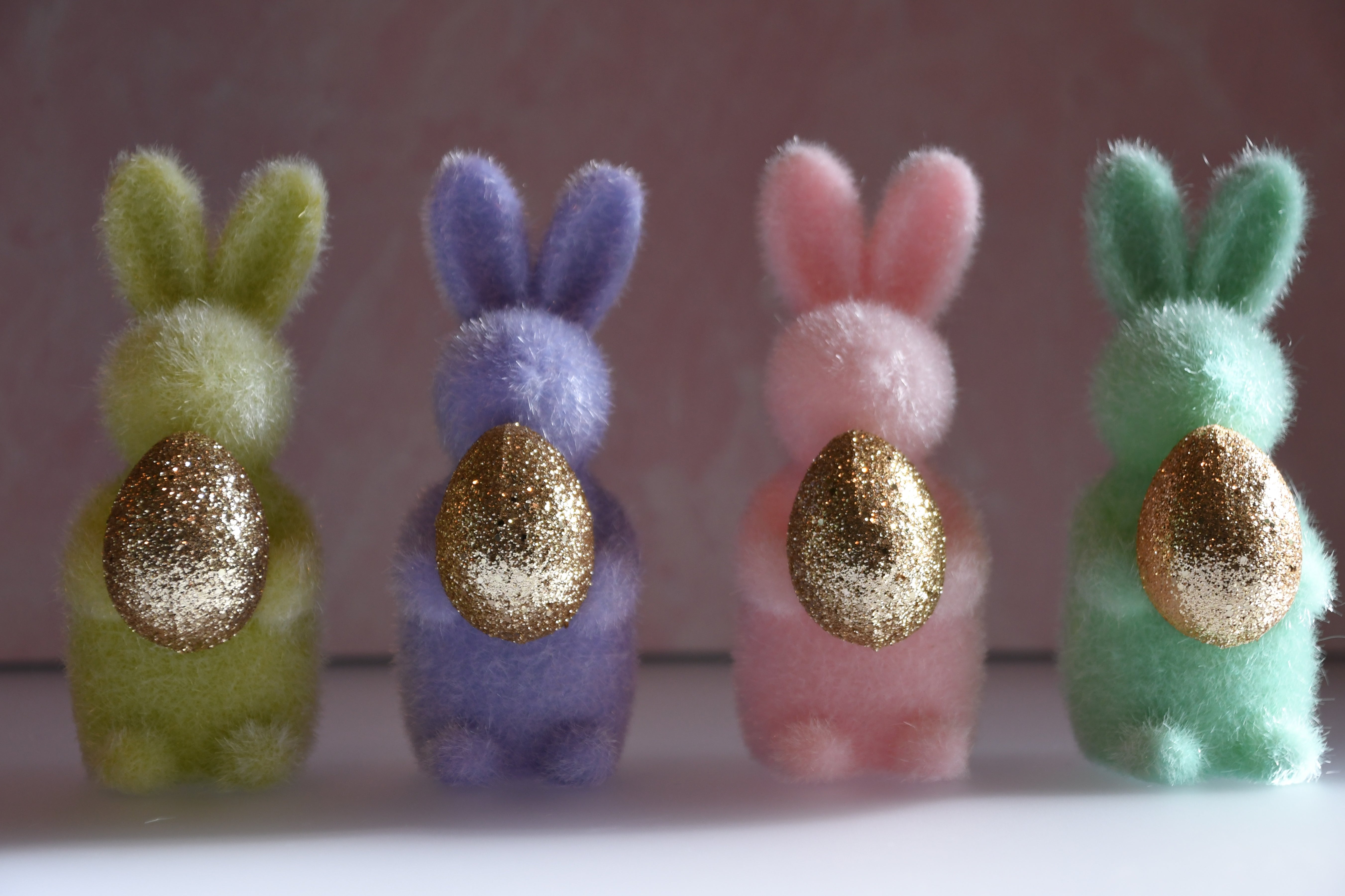 Four colorful felt bunnies with gold eggs on a white surface and light pink background