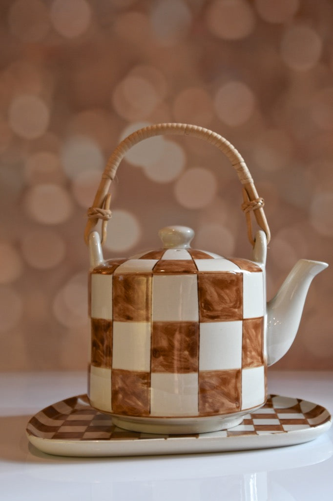 Checkered ceramic teapot with wicker handle on a matching tray against a blurred background