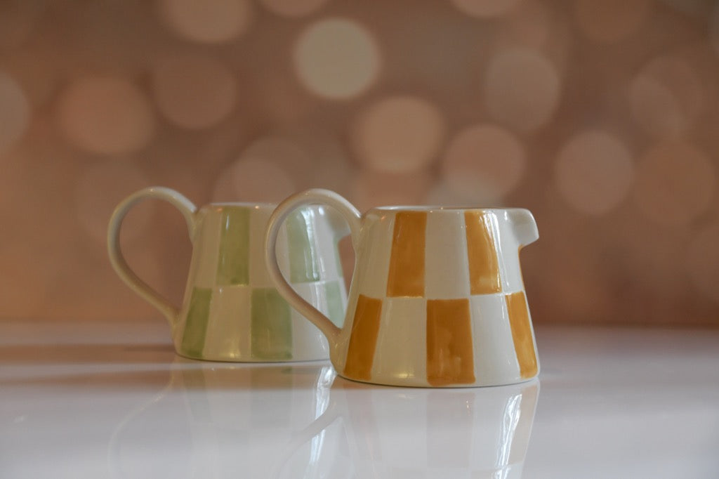 Two ceramic pitchers with checkered patterns on a reflective surface with a blurred bokeh background.