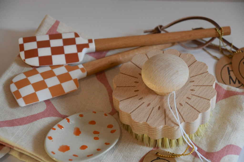 Set of kitchen utensils including checkered spatulas, a wooden brush, and a polka dot dish on a patterned cloth.