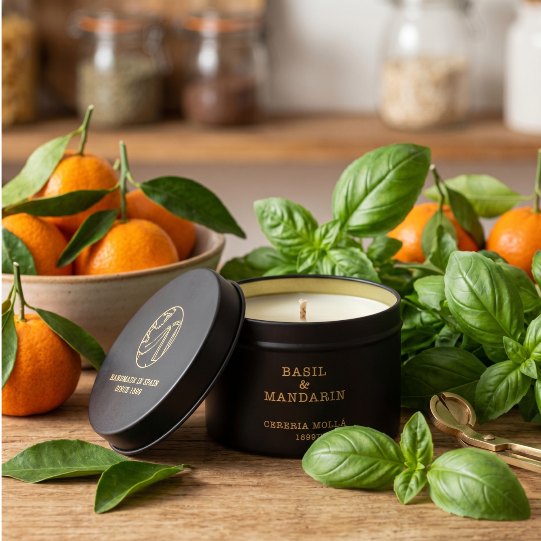 Basil and Mandarin candle in a black tin surrounded by basil leaves and mandarin oranges on a wooden surface.