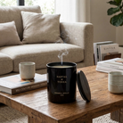 Black candle labeled 'Santal Tonka' on a wooden coffee table with a beige sofa and books in the background.
