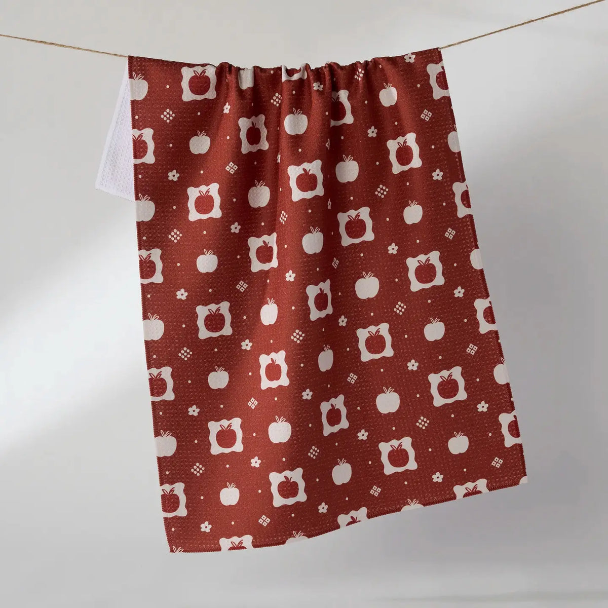 A terracotta red kitchen towel with a white apple and pumpkin pattern, hanging from a string against a plain white wall.