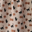 A close-up of a fabric surface with a repeating pattern of black cat silhouettes and brown/orange autumn leaves on a light beige background.