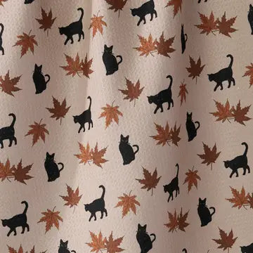 A close-up of a fabric surface with a repeating pattern of black cat silhouettes and brown/orange autumn leaves on a light beige background.