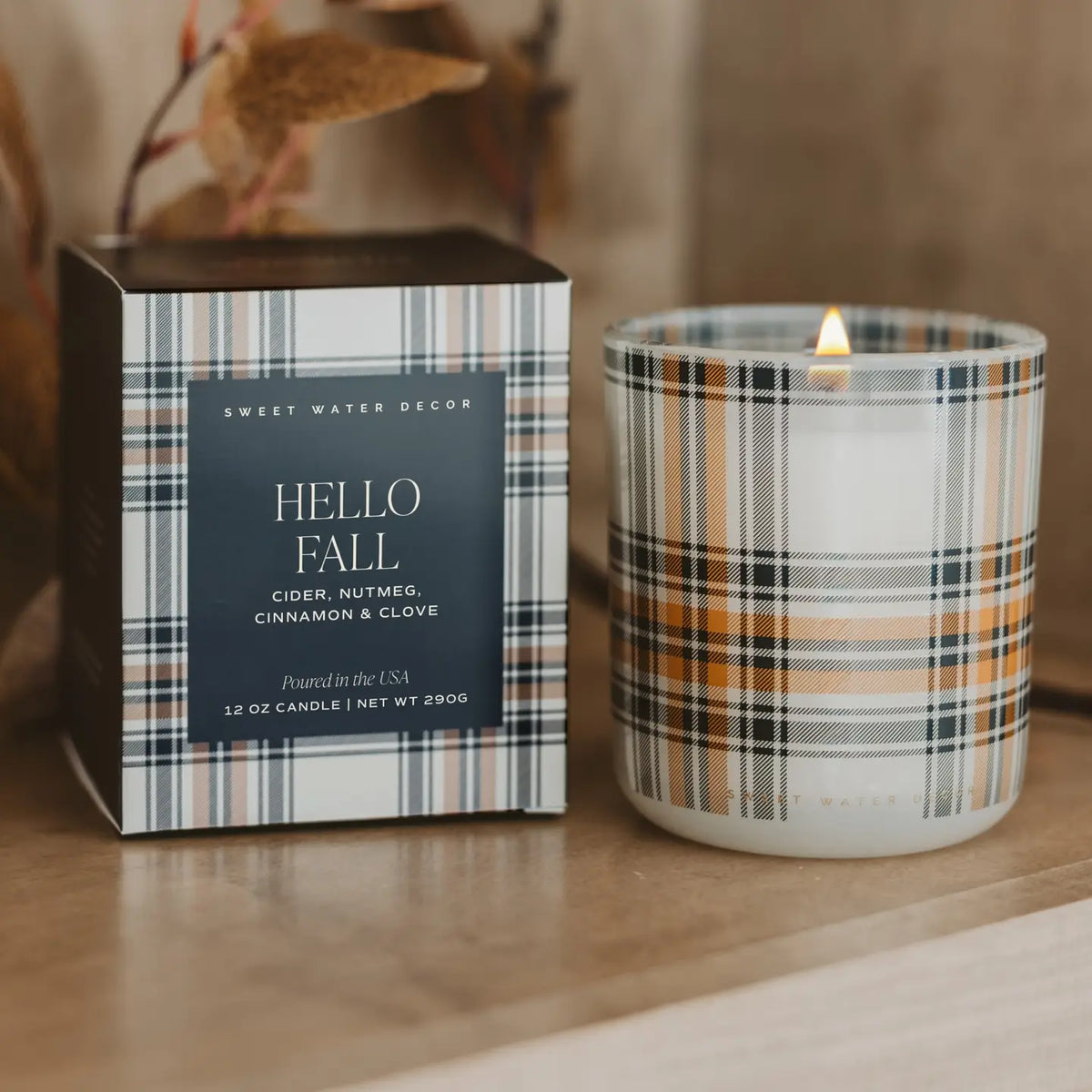 A lit scented candle in a white glass jar with a plaid pattern, standing next to its matching square box on a wooden surface. The plaid features black, gray, white, and warm tan stripes, and the front label reads 'HELLO FALL' by Sweet Water Decor.