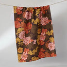 A gathered curtain panel with a dense floral pattern in pink, orange, yellow, and red on a dark brown background, hanging from a thin cord against a light gray wall.