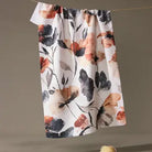A white kitchen towel with a watercolor-style floral print in orange, black, and gray, hanging from a string against a neutral gray background.