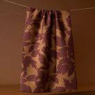 A single curtain panel hanging from a rope against a warm brown wall, featuring a textured waffle-weave pattern of dark plum purple and tan leaf motifs.