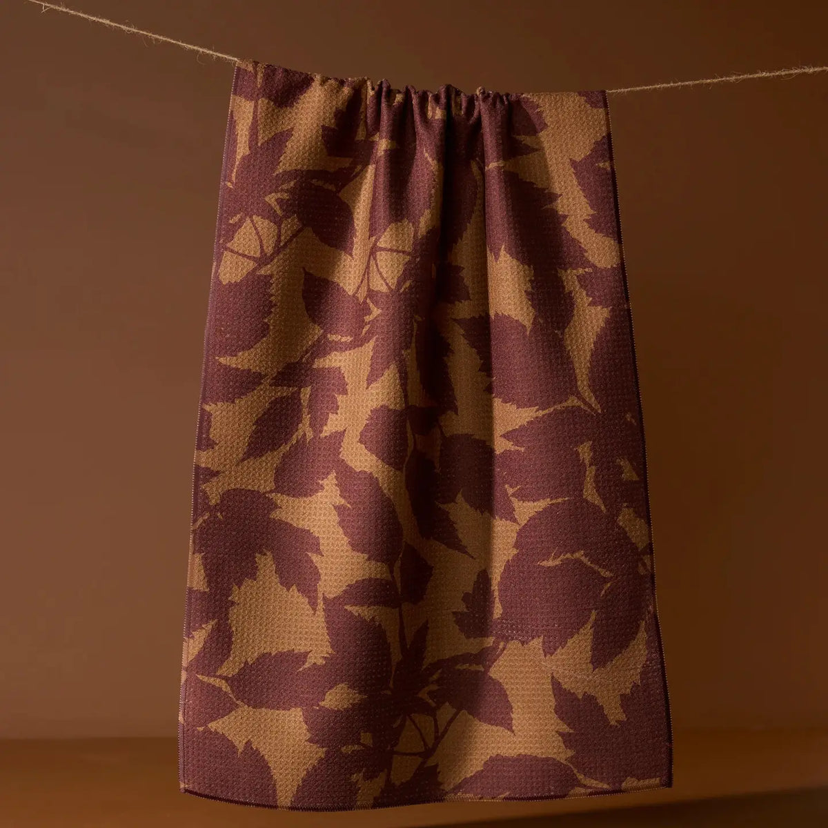 A single curtain panel hanging from a rope against a warm brown wall, featuring a textured waffle-weave pattern of dark plum purple and tan leaf motifs.